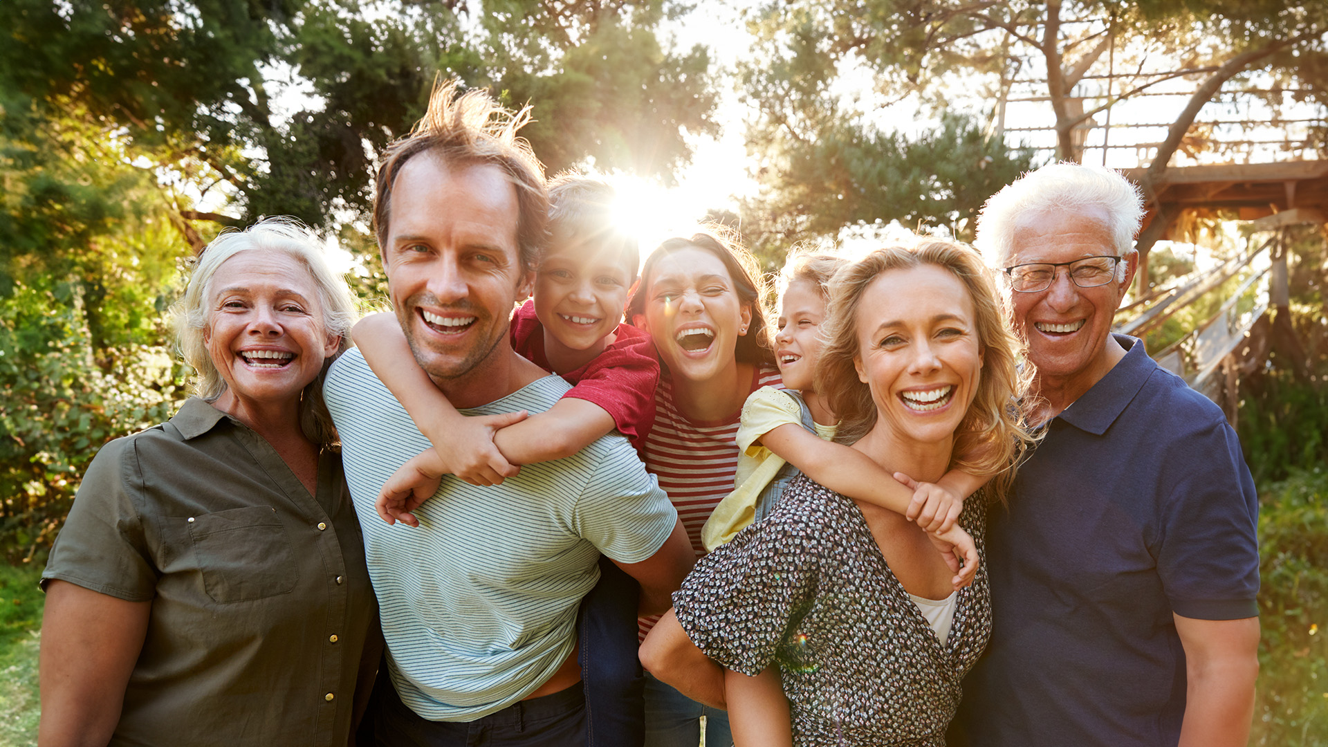 The image shows a group of people, including adults and children, posing together outdoors during daylight with a sunny sky, smiling towards the camera, capturing a moment of familial joy.