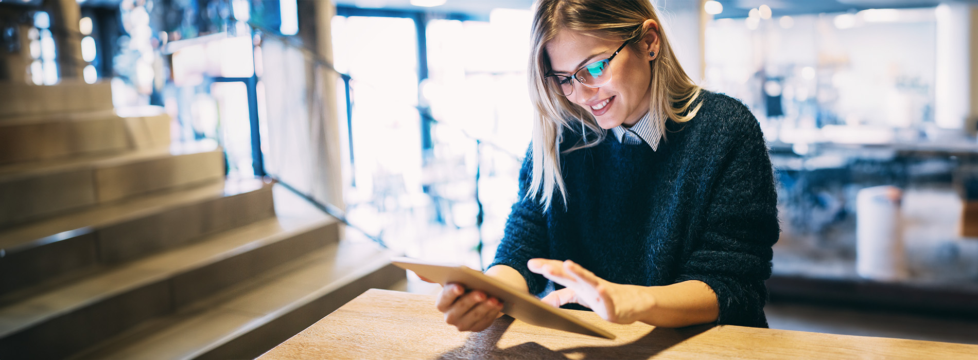 A woman seated at a table inside a store, holding a tablet with her hand, looking up with a smile.