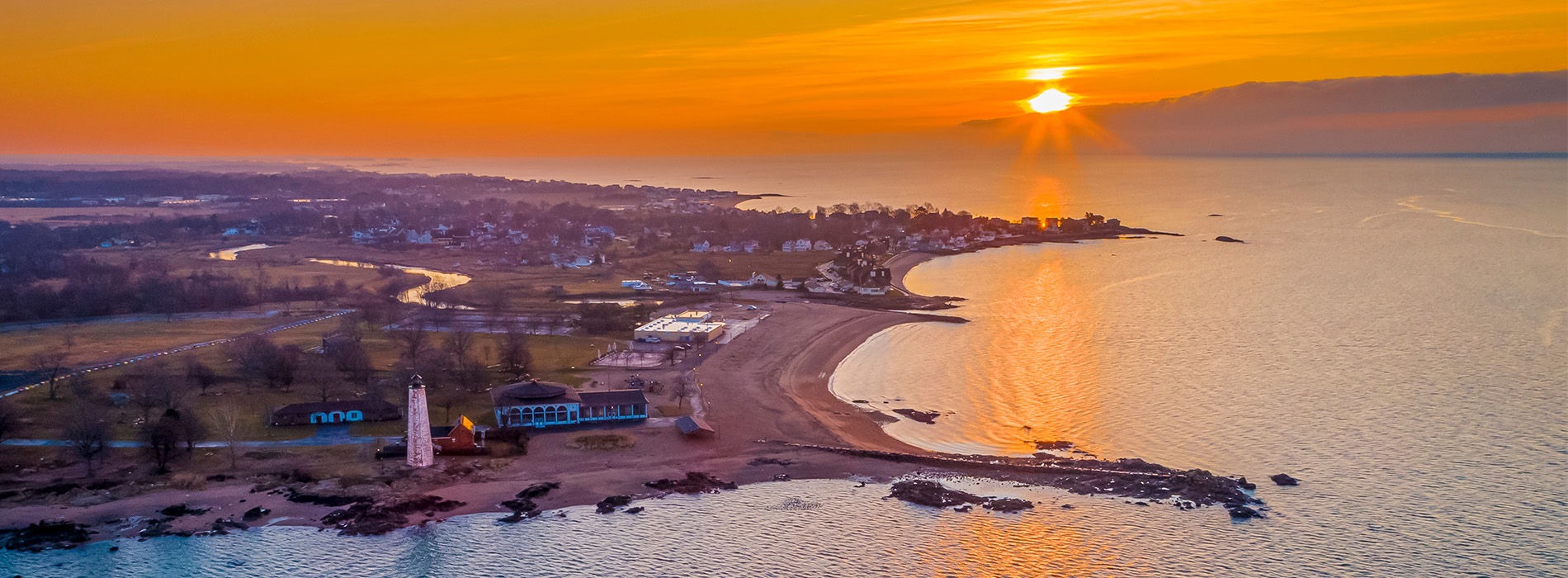 The image shows a sunset over a calm body of water with a beach on the left side, where the sky transitions from warm hues near the horizon to cooler tones at the top, and the reflection of the sun creates a golden glow on the water s surface.