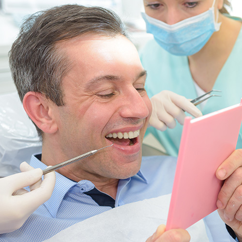 A man sitting in a dental chair with a broad smile, receiving dental treatment from a woman wearing a surgical mask and holding a pink tablet.