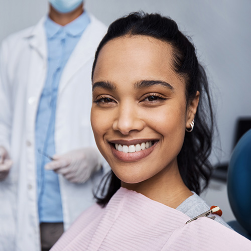 The image features a woman with a radiant smile, seated in a dental chair, wearing a protective face mask, with a dental professional standing behind her, all set within a dental office environment.