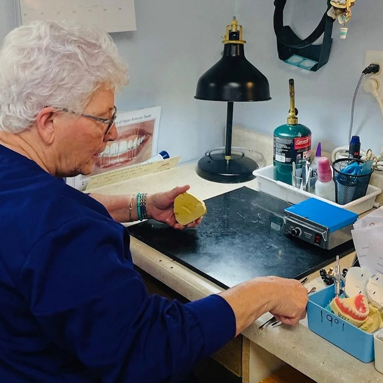 The image shows an older adult woman seated at a dental office workstation, holding a small yellow object, possibly a dental tool, with her right hand, while working on a piece of paper with a pen.