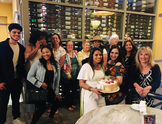 A group of people celebrating at a restaurant, with a cake on the table and wine bottles in the background.