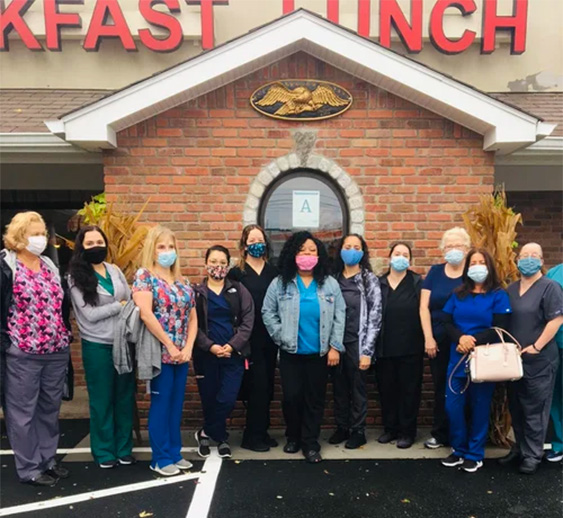 The image shows a group of healthcare professionals wearing face masks standing outside a restaurant with a sign indicating  BREAKFAST LUNCH  and a logo on the wall behind them.