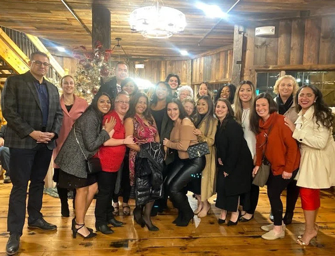 The image shows a group of people posing together indoors, with some individuals smiling at the camera and others looking towards something off-frame. They are standing in front of a rustic wooden wall with a Christmas tree visible in the background.