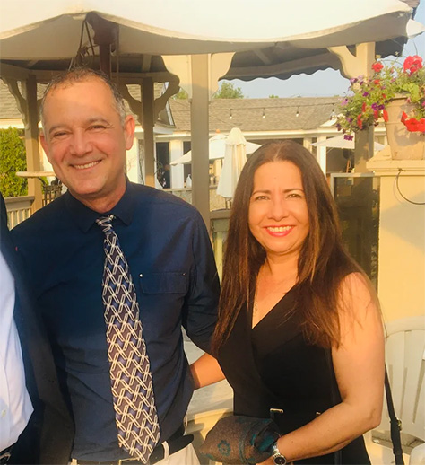 The image shows two adults posing together outdoors at an evening event, with a man wearing a blue shirt and tie standing next to a woman who appears to be laughing. They are smiling and seem to be enjoying themselves.