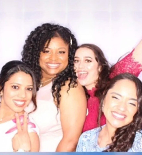 The image shows a group of people posing together at an event, with a woman in the center smiling towards the camera while others stand around her, some making hand gestures. They appear to be dressed for a formal occasion.