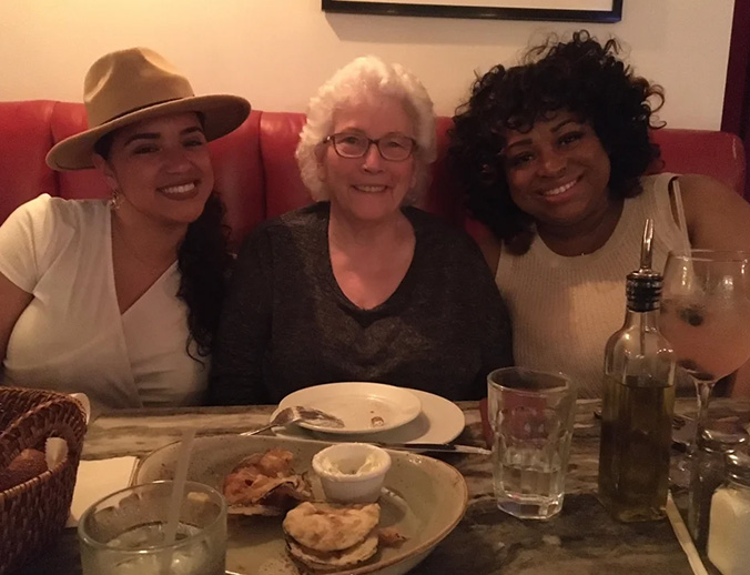 The image shows three individuals posing at a dining table with food, drinks, and restaurant decor visible in the background.