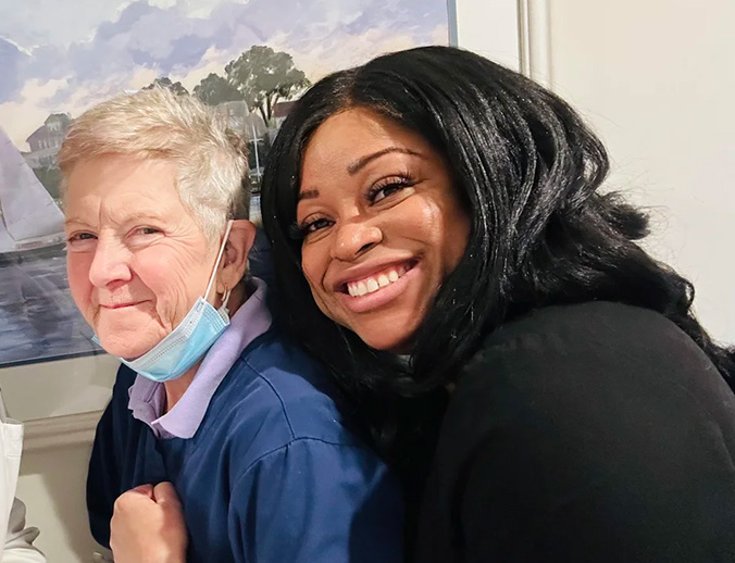 Two individuals, likely medical professionals, posing together with smiles; one wearing a white mask and the other a blue surgical gown.