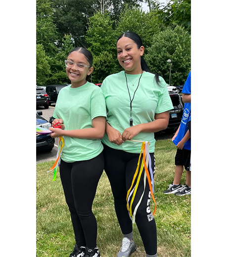 The image shows two individuals wearing matching green t-shirts with a lanyard around their necks, posing for a photo outdoors during daylight hours.