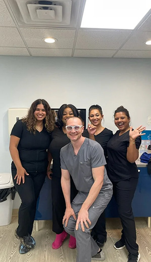 A group of five individuals, including two standing and three sitting, posing for a photo in an office setting with a dental chair visible in the background.