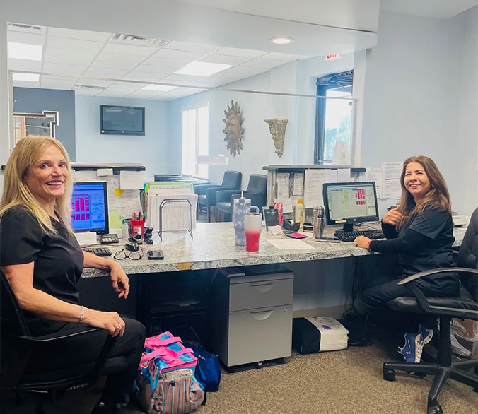 The image shows four individuals seated at an office desk, with two women on the left side of the frame and two more on the right, all engaged in work activities.
