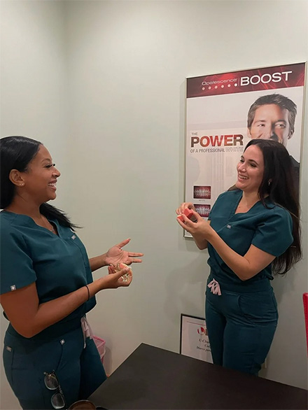 Two women in scrubs standing side by side and smiling at each other in an office setting.