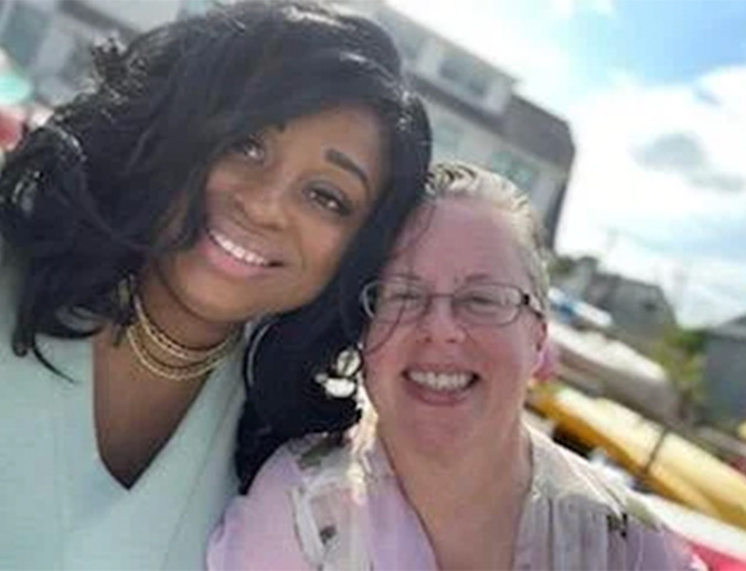 The image shows two individuals posing together  one is a woman with long hair wearing a black top and a necklace, smiling at the camera, while the other appears to be an older individual, also smiling and looking towards the camera. They are outdoors during the daytime under clear skies.