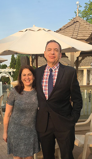 In the image, there are two individuals standing outdoors at an event, with one person wearing a suit and tie, both posing for the camera.