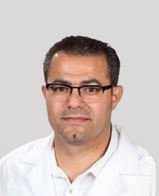 The image shows a man wearing glasses with a beard, smiling at the camera. He has dark hair, and he s dressed in a white shirt with a collar, suggesting a professional setting.