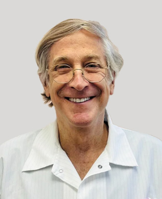 The image shows a man smiling at the camera, wearing glasses and a white shirt with a name tag, standing against a plain background.