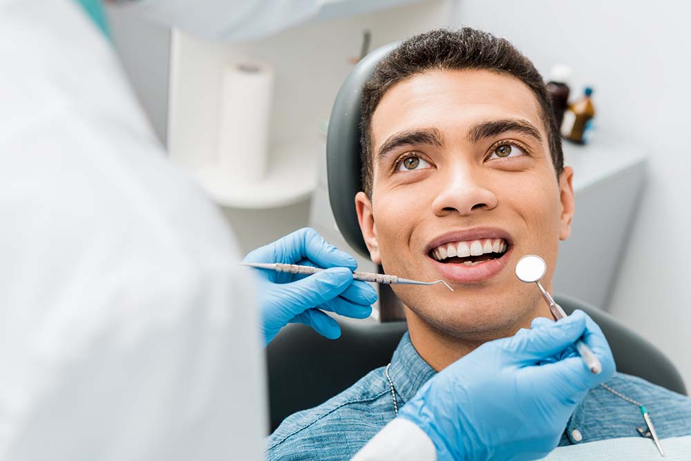 The image shows a person in a dental chair receiving treatment from a dentist, with the patient smiling and looking at the camera, while the dentist is focused on the procedure.