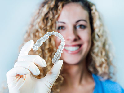 A woman is holding up a clear plastic dental retainer with her left hand, smiling at the camera.