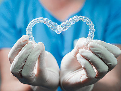 A person s hands gently cradling a heart-shaped transparent object, which appears to be a dental retainer or mouthguard, set against a blue background.