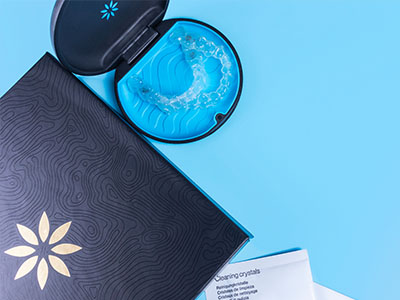 The image shows a cosmetic product, specifically a jar with a lid and a patterned box on a blue surface, alongside a blue item resembling a toothbrush cleaner.