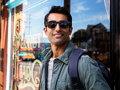 A man poses confidently on a city street, leaning against a window with sunglasses, wearing a backpack and casual attire.