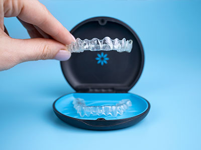 The image shows a hand holding an open box with a clear plastic tray containing a set of transparent dental aligners, which are used for orthodontic purposes.