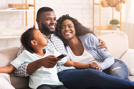A family of four sitting on a couch with a man holding a child, smiling at the camera, while a woman embraces them both.
