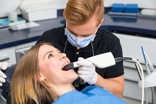 The image shows a dental hygienist performing a cleaning procedure on a patient s teeth using an electric toothbrush, with the patient seated in a dental chair and wearing protective eyewear.