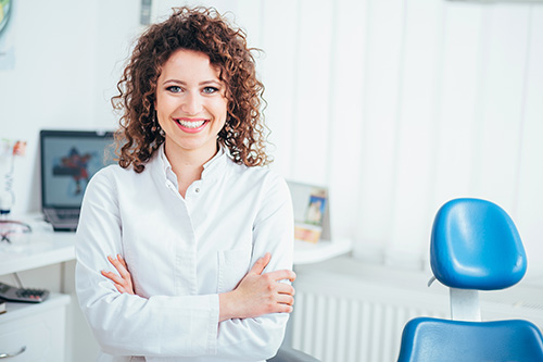 A woman with curly hair stands confidently in an office setting, smiling at the camera, dressed in a white shirt and black pants.
