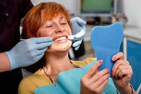 A woman with red hair is seated in a dental chair, smiling at the camera while holding a blue mouthguard, with a dentist adjusting her teeth.