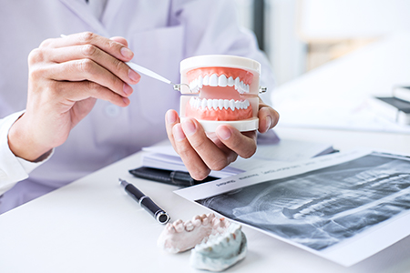 A split-screen photograph showing two different moments  on the left, a dental professional s hands holding a model of a human mouth with an open jaw  on the right, another dental professional seated at a desk with various instruments and materials in front of them, examining a dental model with a magnifying glass.