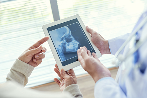 The image depicts two individuals examining a tablet displaying an X-ray of a human skull, with a medical professional holding the tablet and viewing the X-ray alongside another person.