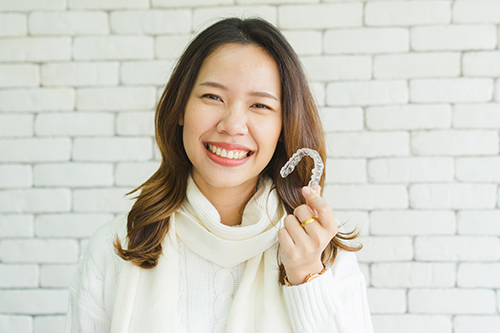 A woman with dark hair smiling at the camera, holding a toothbrush, posing against a brick wall background.