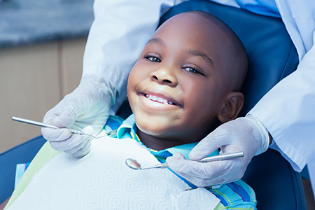 A young boy is sitting in a dentist s chair, smiling at the camera while receiving dental care, with a dental professional standing behind him wearing protective gloves and a face mask.