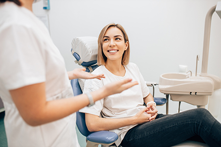 The image depicts a dental office setting with a female patient seated in a chair, smiling at the camera, while a dental professional stands behind her, also facing the camera.